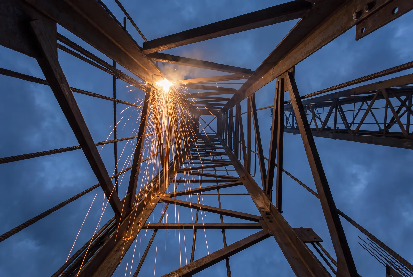 Looking up a steel column cage at dusk with welding sparks falling toward the camera